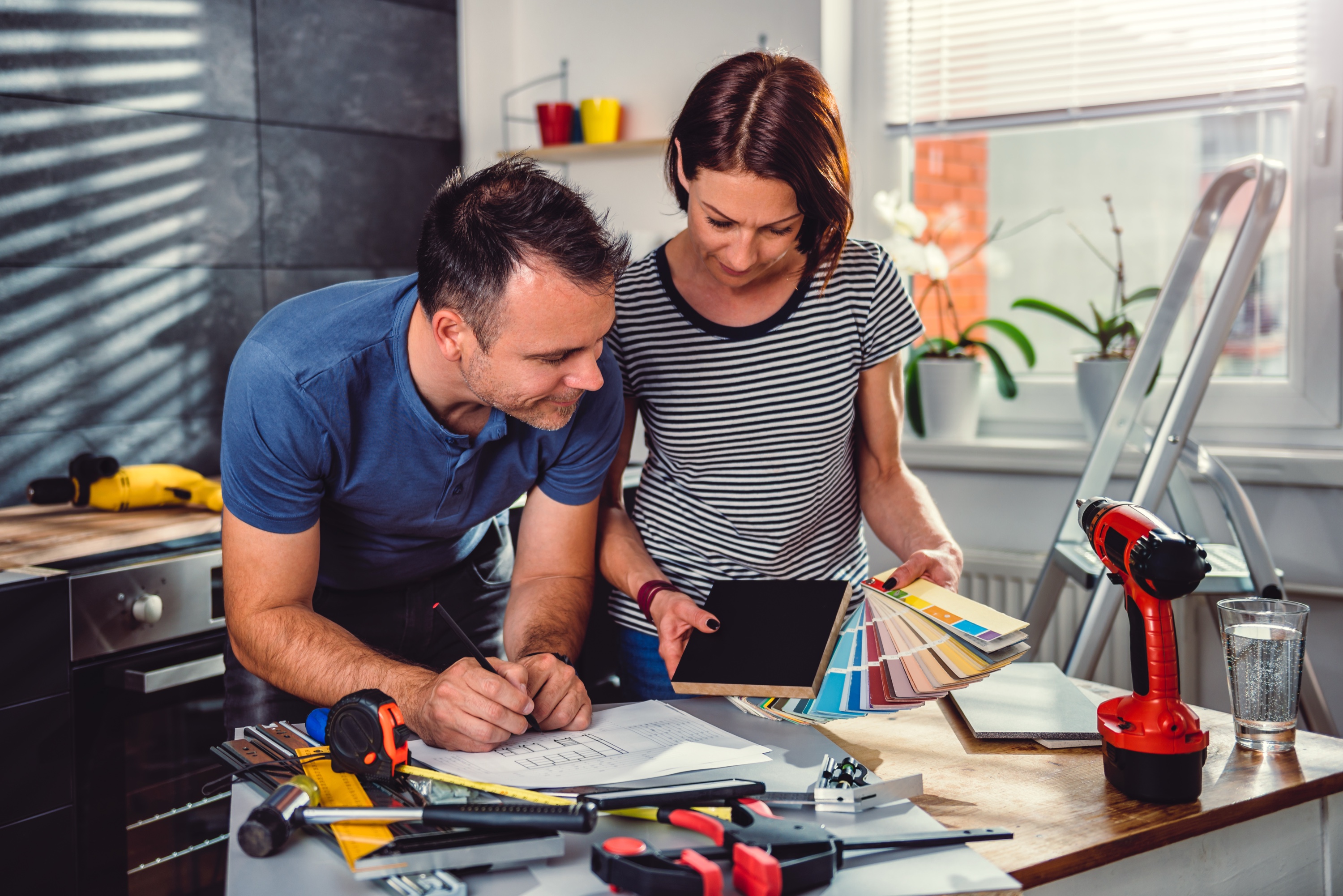 Couple at a kitchen island reviewing renovation plans with paint color swatches, a drill on the counter and a ladder in the background