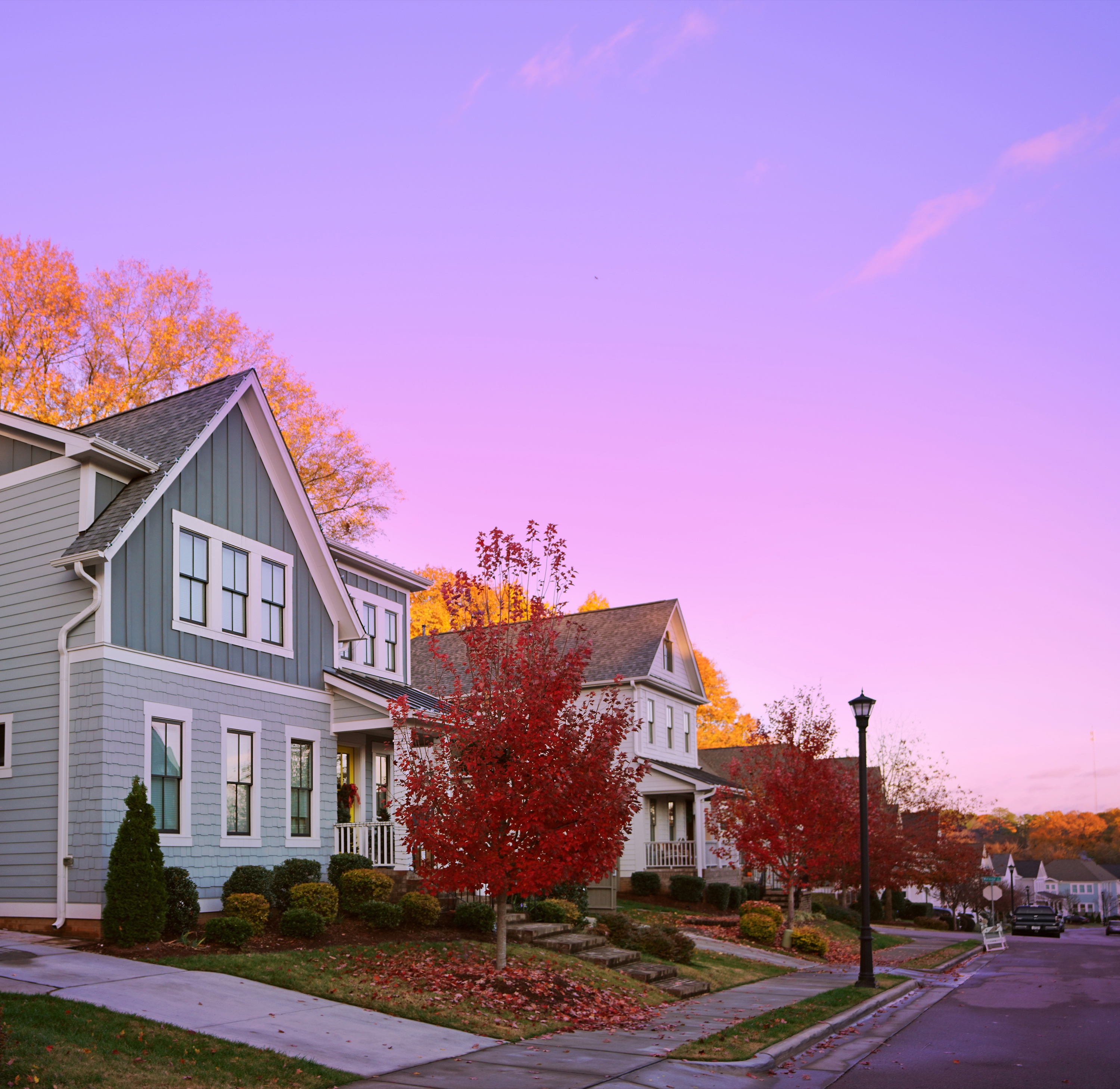 New-build suburban street at twilight with a blue board-and-batten home and a white modern farmhouse with a front porch, lamp posts along the sidewalk