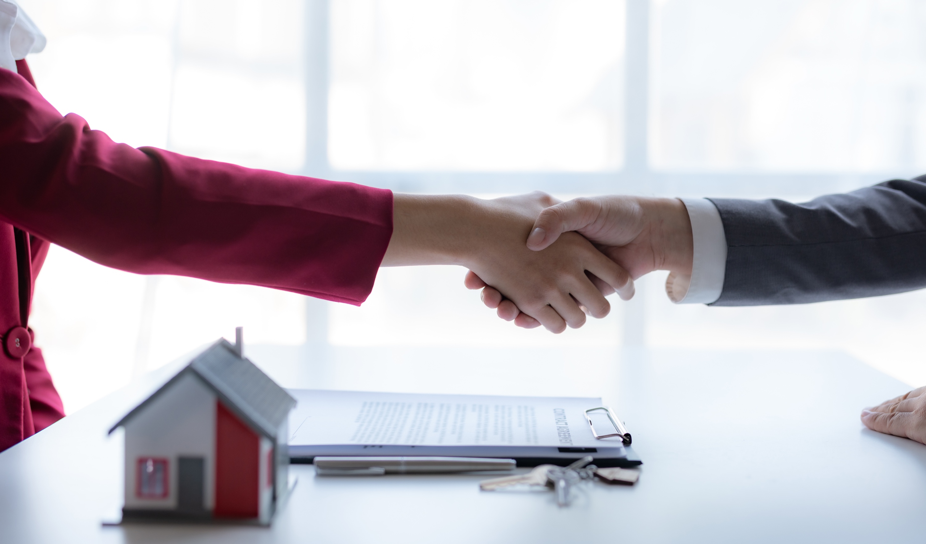 Two people shaking hands over a desk with a small red-roof house model, a contract, a pen and keys