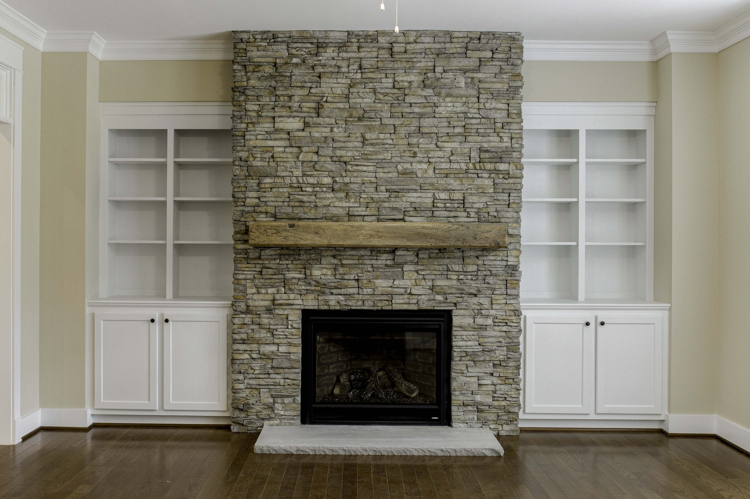 Empty traditional living room with a stacked-stone fireplace, rustic wood mantel and symmetrical white built-in bookcases